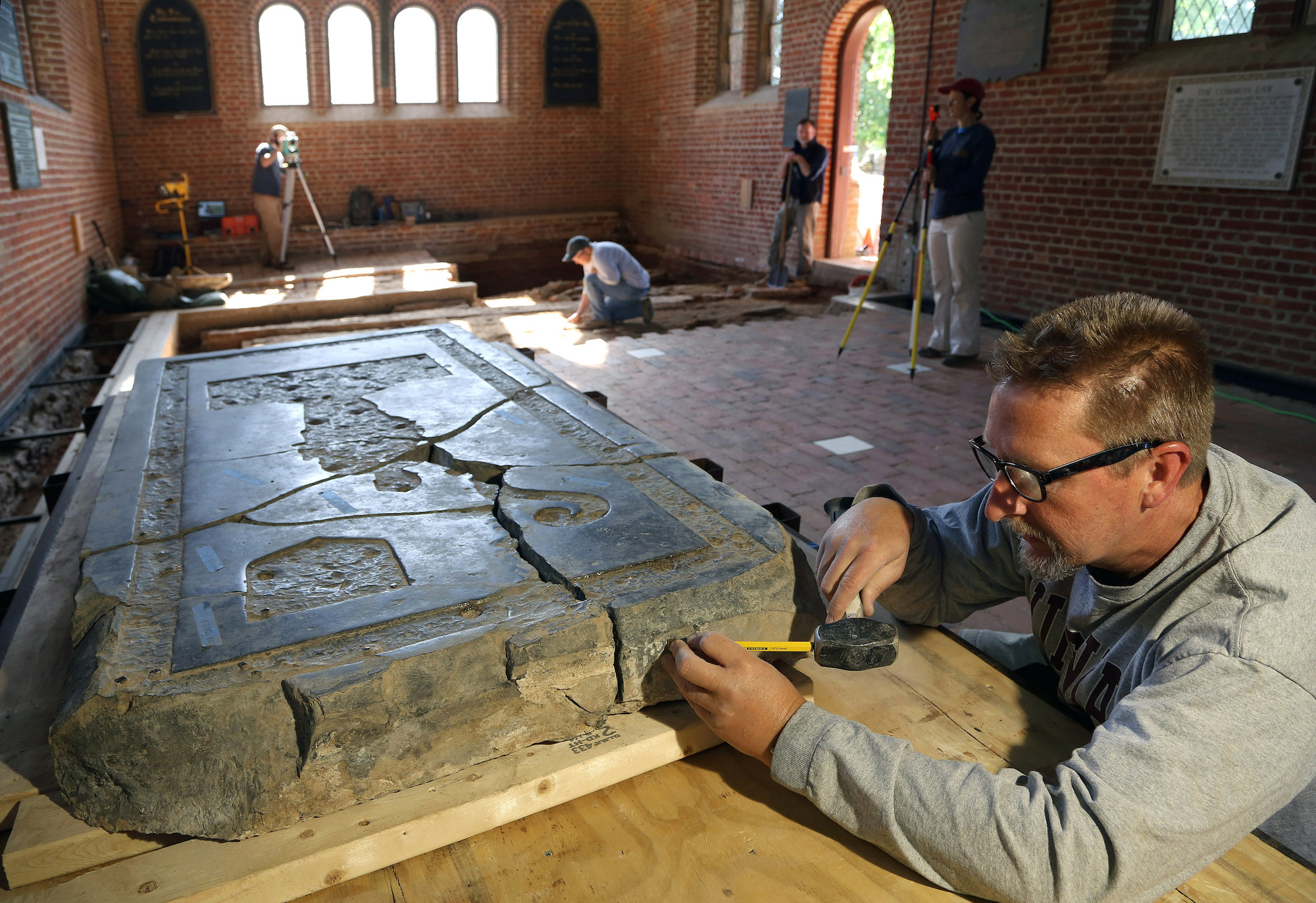 Senior Conservator Dan Gamble works on conservation of the tombstone while archaeologists excavate in the background