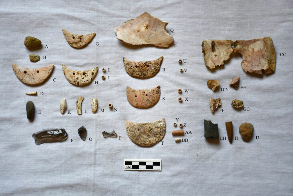 Grave goods from the woman's burial, including mascaras (C, G, H, R, S, T), spondylus pendants (Q, CC), green stone (A, B, E), obsidian (F, O, P), chaquira (I, J, N, U, V, W, X, Y, Z), other beads (K, M, AA, BB), shell and spondylus fragments (L, DD, EE, FF, GG), ceramic fragments (HH, II, JJ), and the crab claw